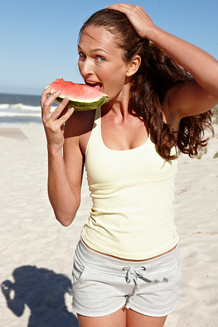 A brunette woman on a beach with a slice of watermelon wearing a light top and shorts