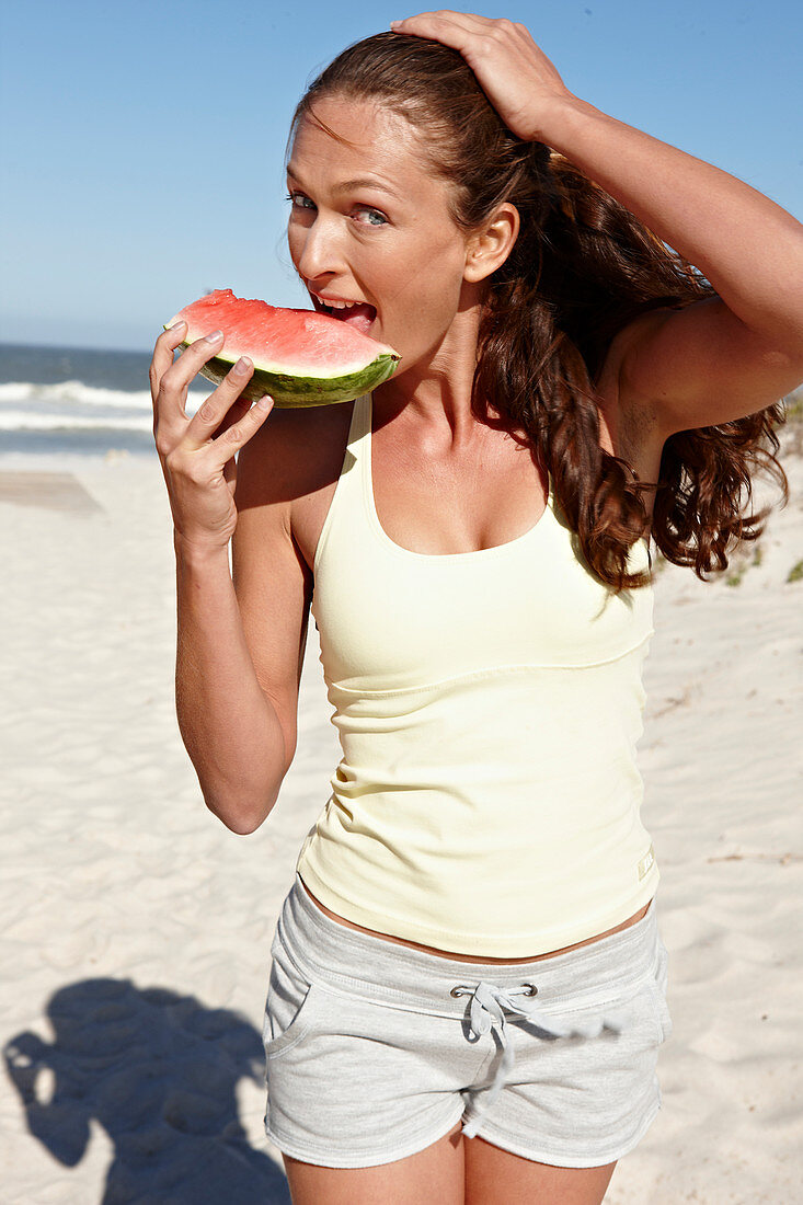 Brünette Frau mit Wassermelone in hellem Top und Shorts am Strand