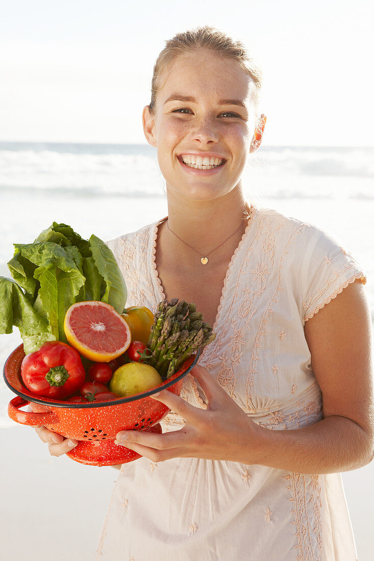 A blonde woman by the sea with a bowl of fruit and vegetables wearing a white dress