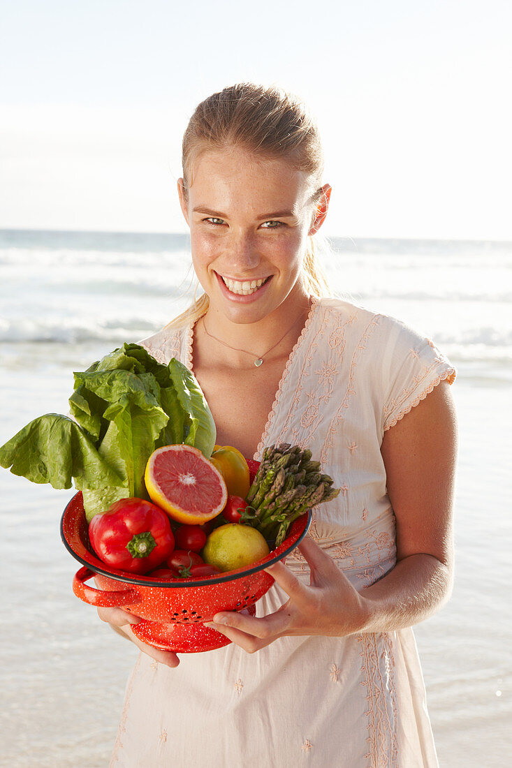 A blonde woman by the sea with a bowl of fruit and vegetables wearing a white dress