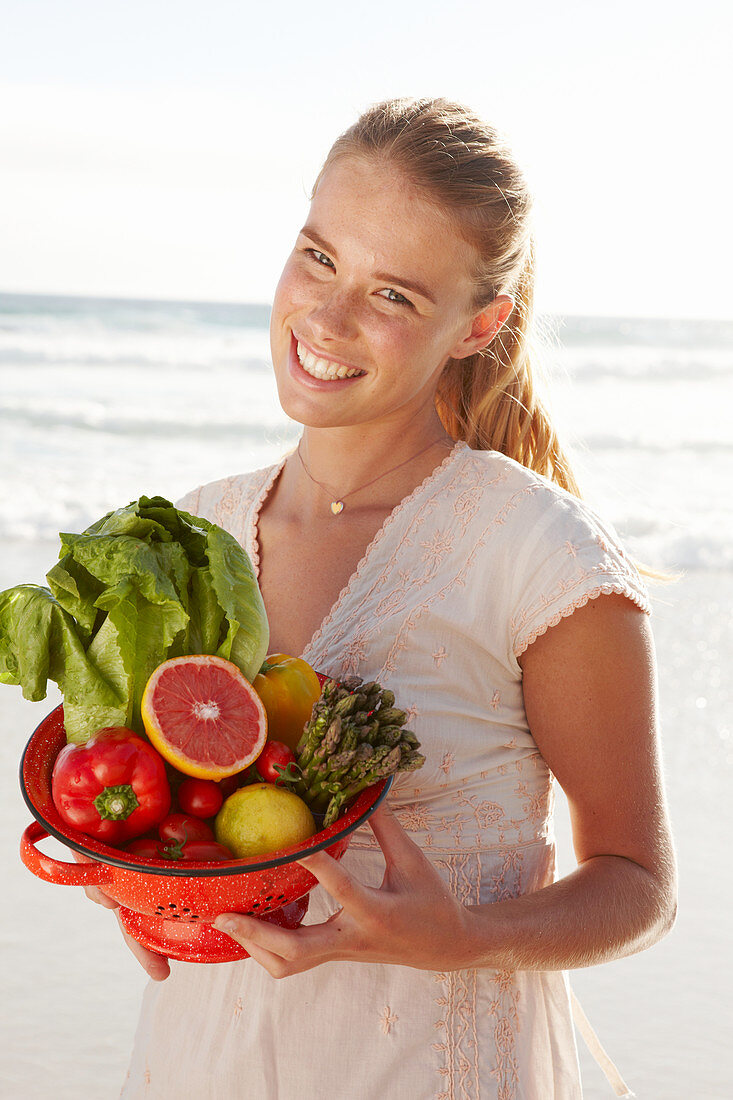 A blonde woman by the sea with a bowl of fruit and vegetables wearing a white dress