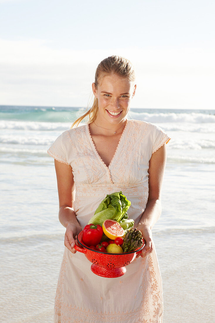 Blonde Frau mit Obst- und Gemüseschale in weißem Kleid am Meer