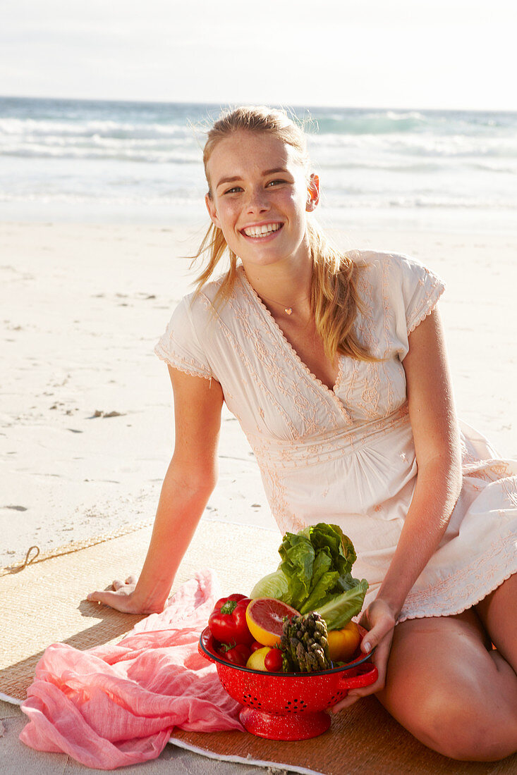 A blonde woman by the sea with a bowl of fruit and vegetables wearing a white dress