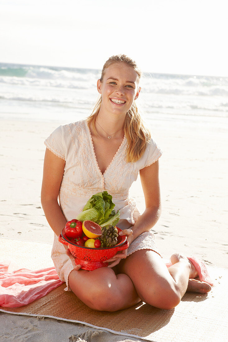 Blonde Frau mit Obst- und Gemüseschale in weißem Kleid am Meer