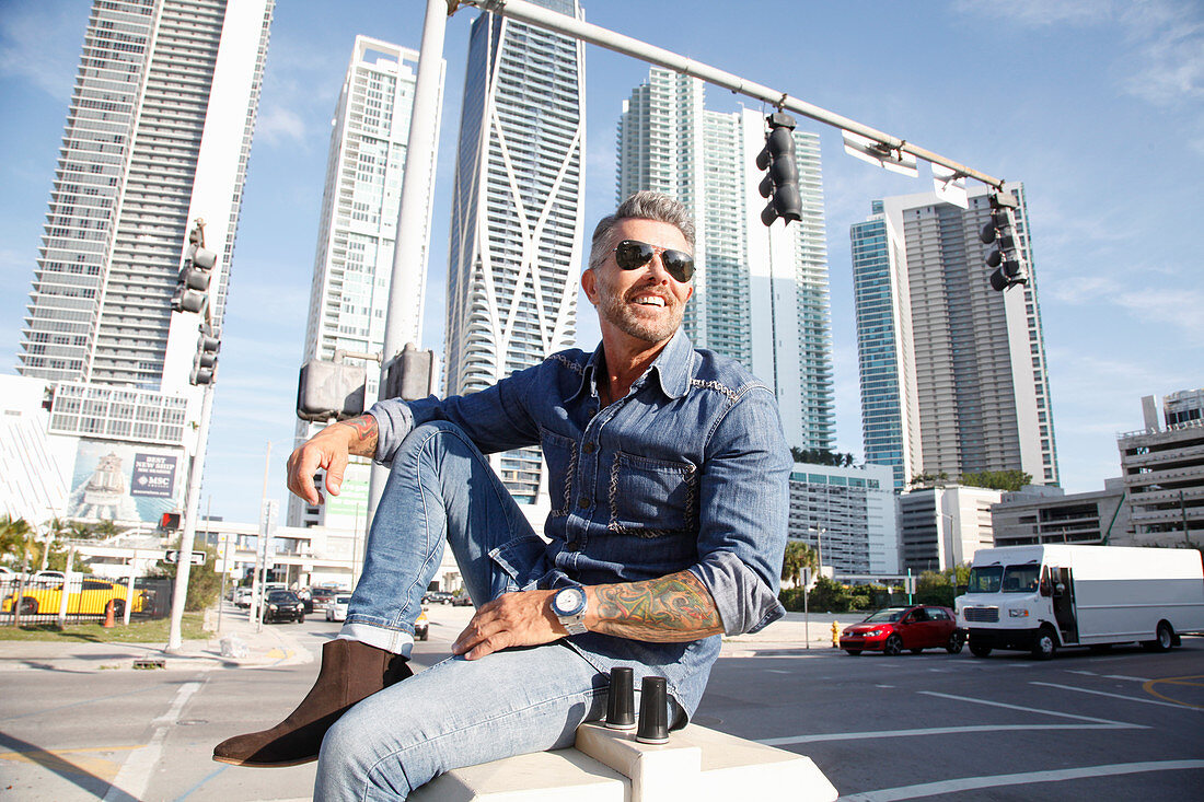 A man with a bread wearing a denim shirt and jeans sitting against a skyline backdrop