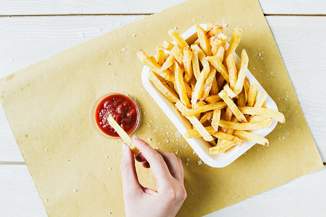 A portion of chips with sea salt, with a hand dipping one into ketchup