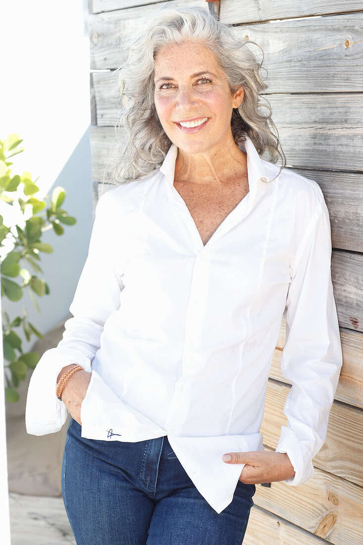 A mature woman wearing a white shirt and jeans standing against a wooden wall