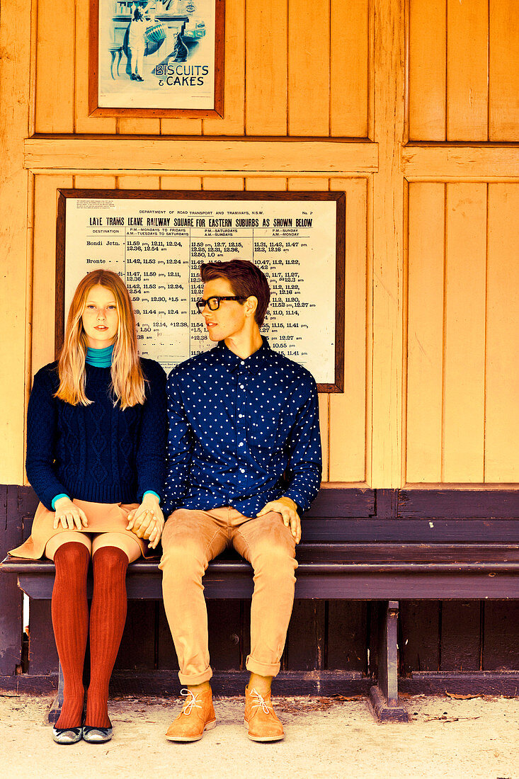 A young couple sitting on a bench holding hands, her wearing a skirt and tights and him wearing a shirt and trousers
