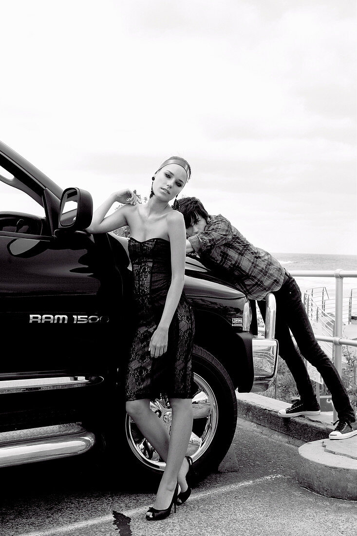 A young woman wearing an off-the-shoulder party dress and a young man wearing a casual outfit standing by a car (black-and-white shot)