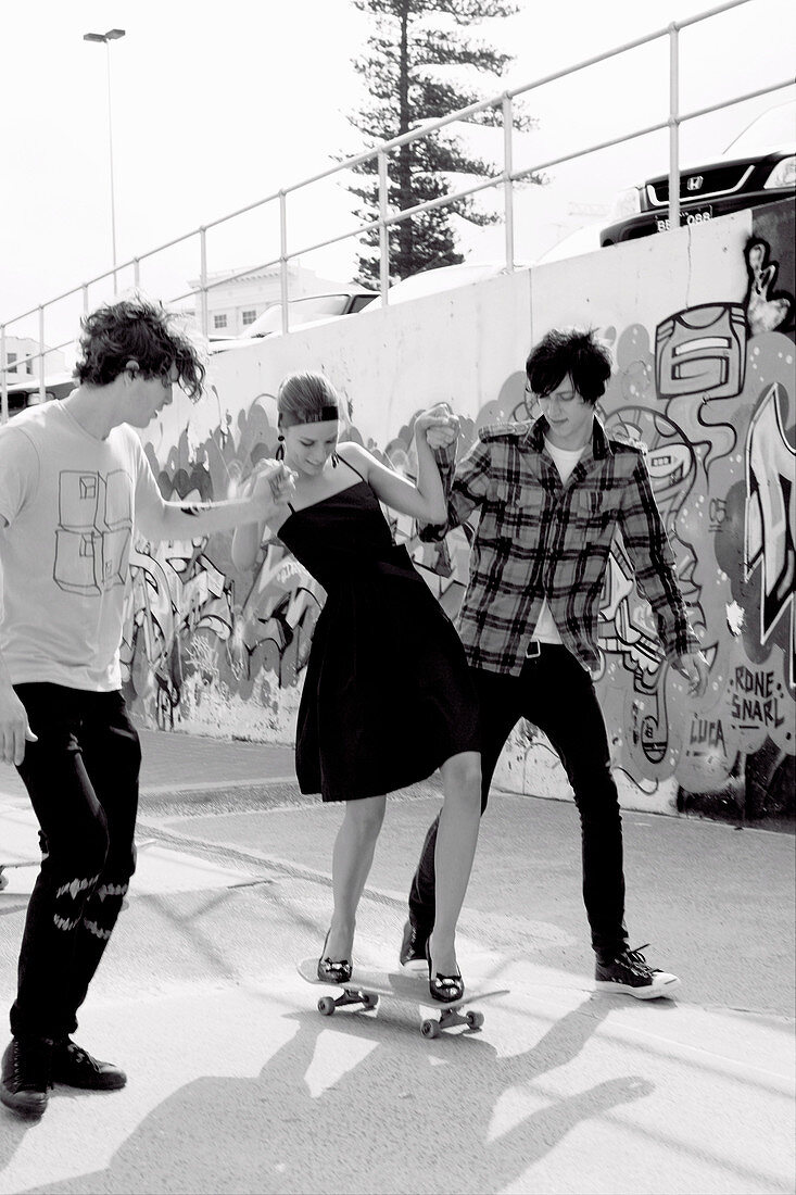 A young woman on a skateboard wearing a party dress being supported by two young men (black-and-white shot)