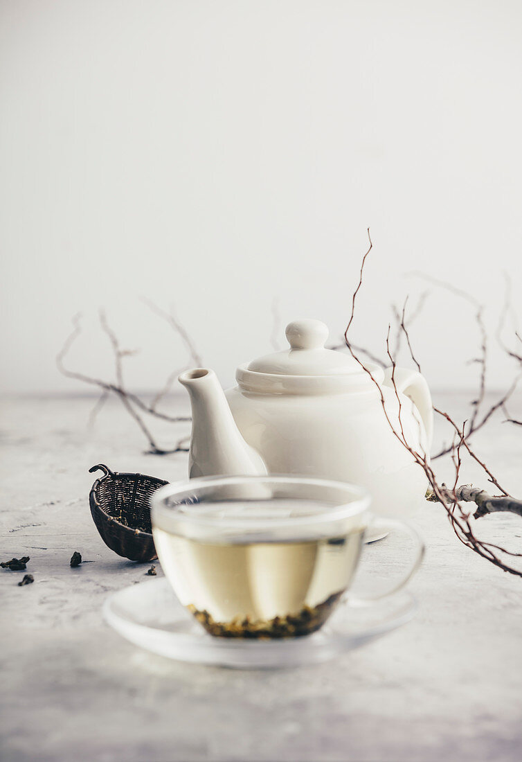 Stillleben mit grünem Tee in Glastasse, Teesieb und weißer Teekanne