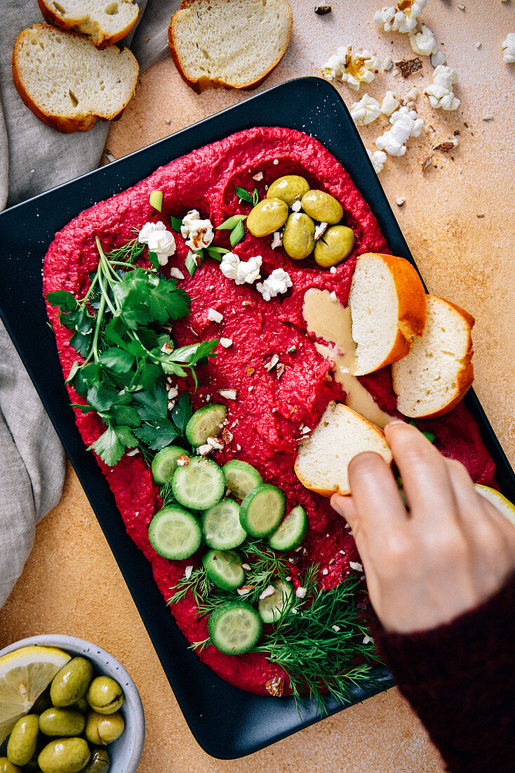 Woman dipping a slice of bread into beet hummus garnished with herbs, cucumber, olives, tahini and lemon