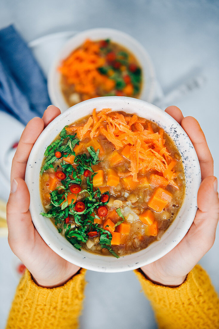 A woman with yellow cardigan holding a bowl of vegan carrot soup with rice, lentils and celeriac