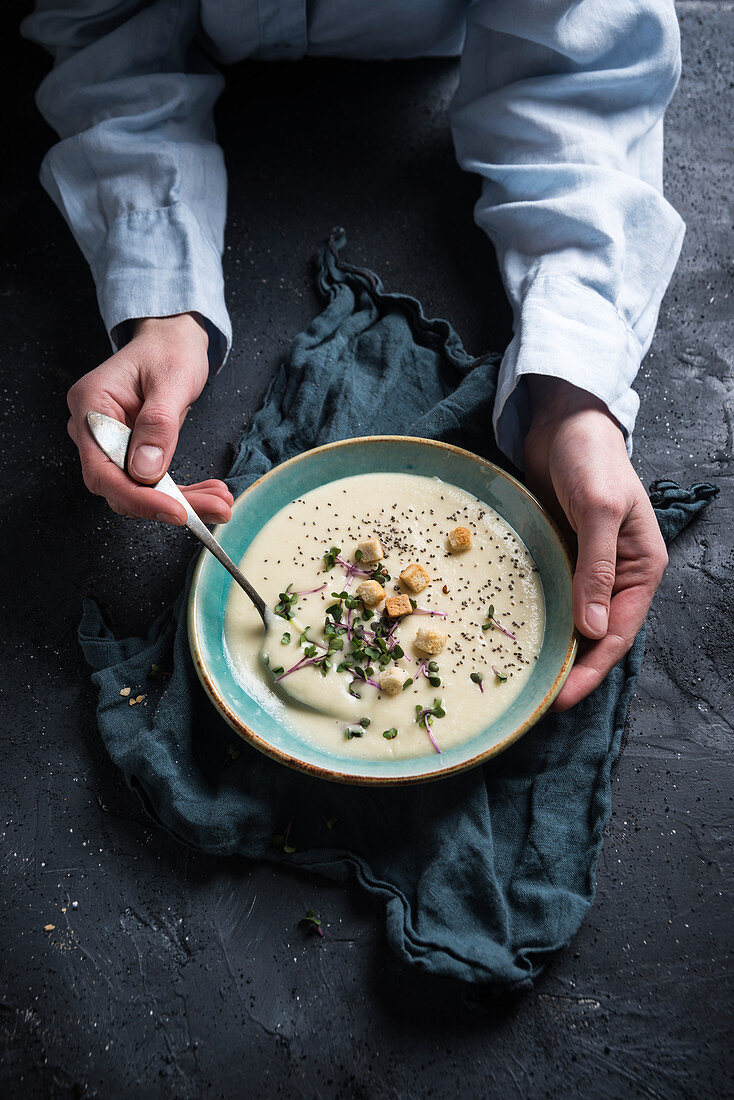 A woman holding a bowl of vegan celeriac soup with croutons, chia seeds and radish cress