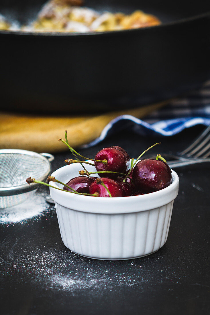 A bowl of fresh cherries next to a sieve of icing sugar