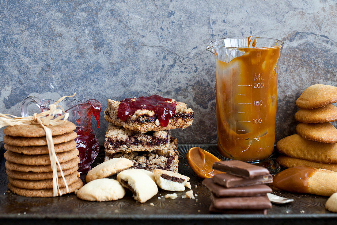 Various cookies stacked on an antique baking sheet, with jam and caramel sauce