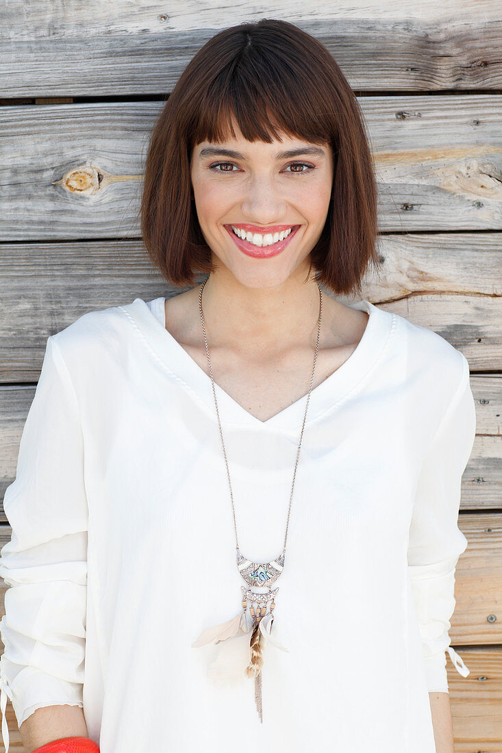 A brunette woman wearing a white long-sleeved blouse standing in front of a wooden wall