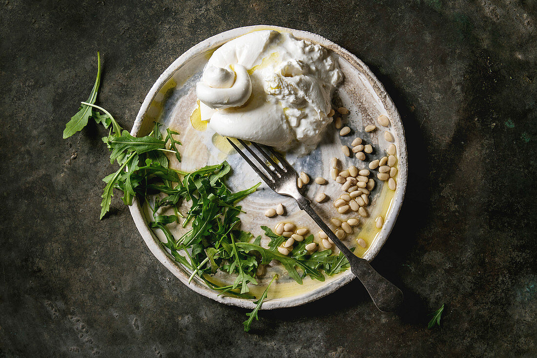 Sliced Italian burrata cheese, fresh arugula salad, pine nuts and olive oil in white ceramic plate