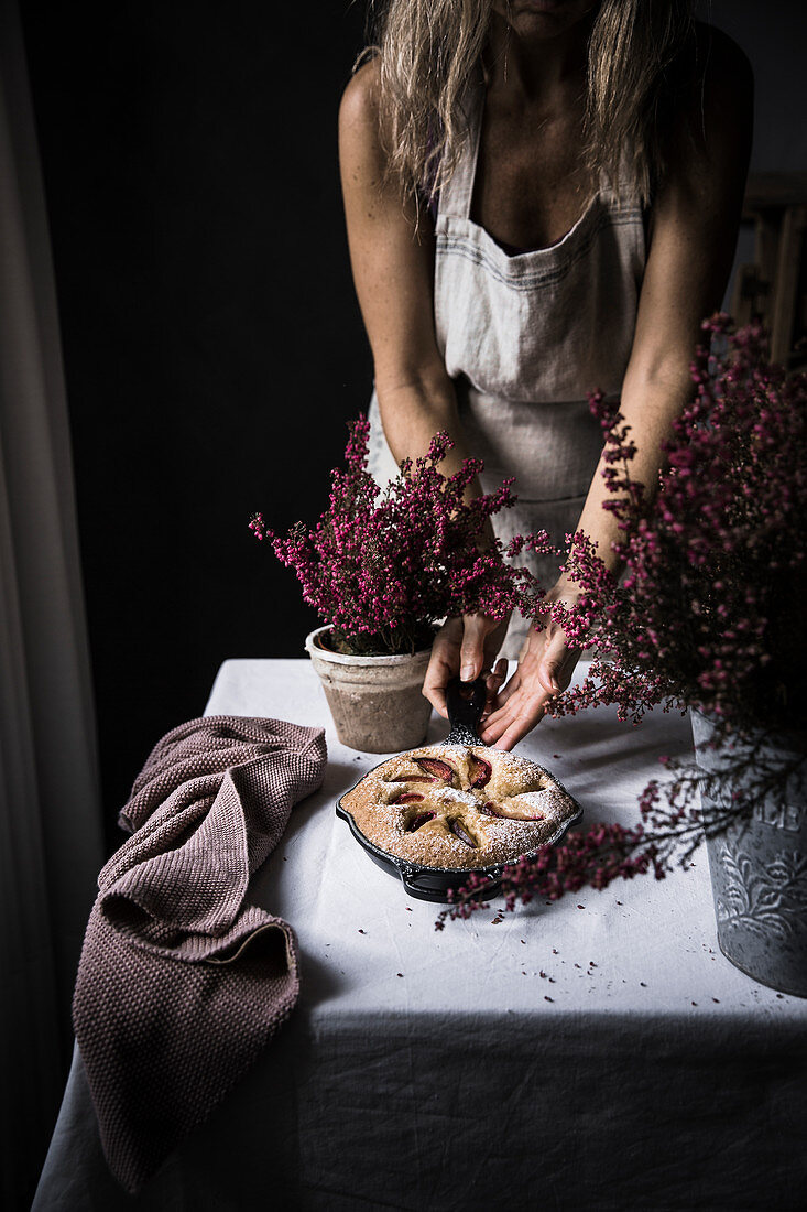 Woman cook in apron cutting plum cake on table with flowers