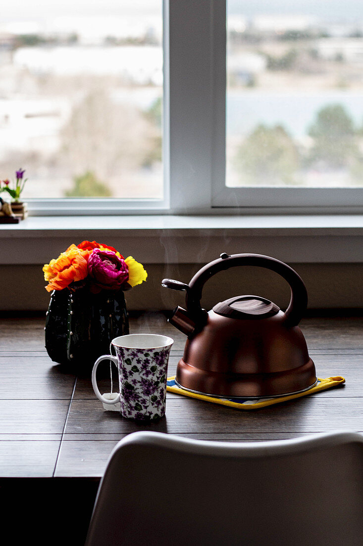 Teacup, tea kettle and a flower vase on a table in front of a window