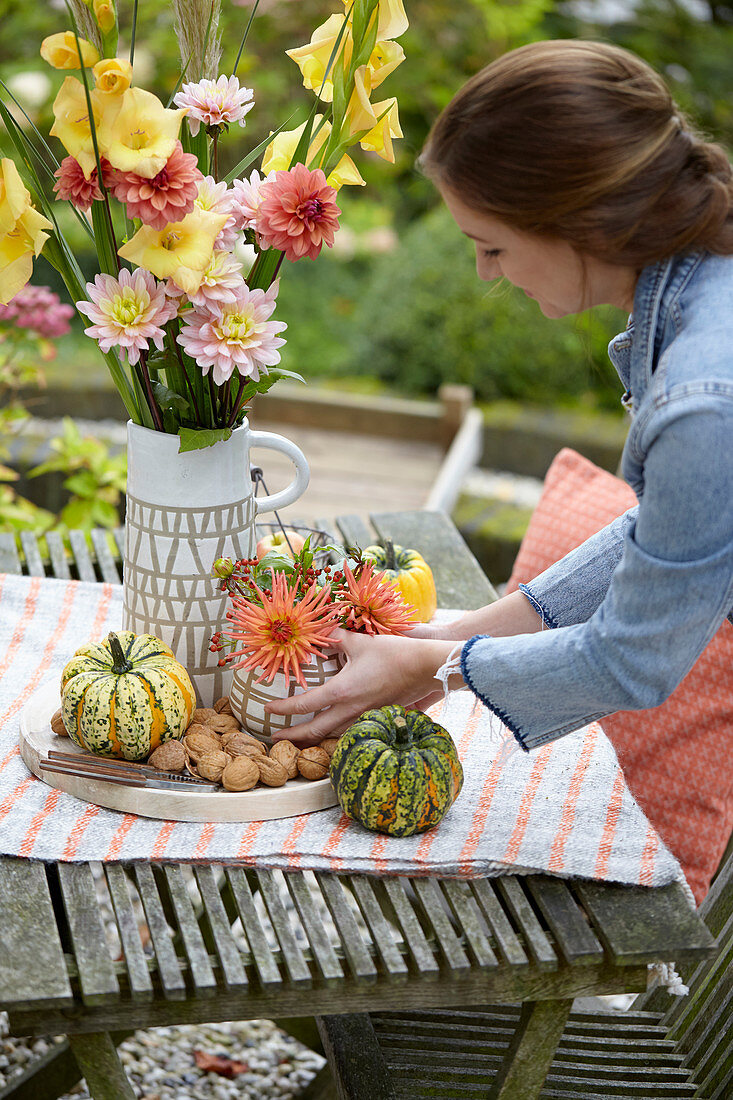 Frau dekoriert Gartentisch mit Dahlien, Gladiolen und Kürbissen