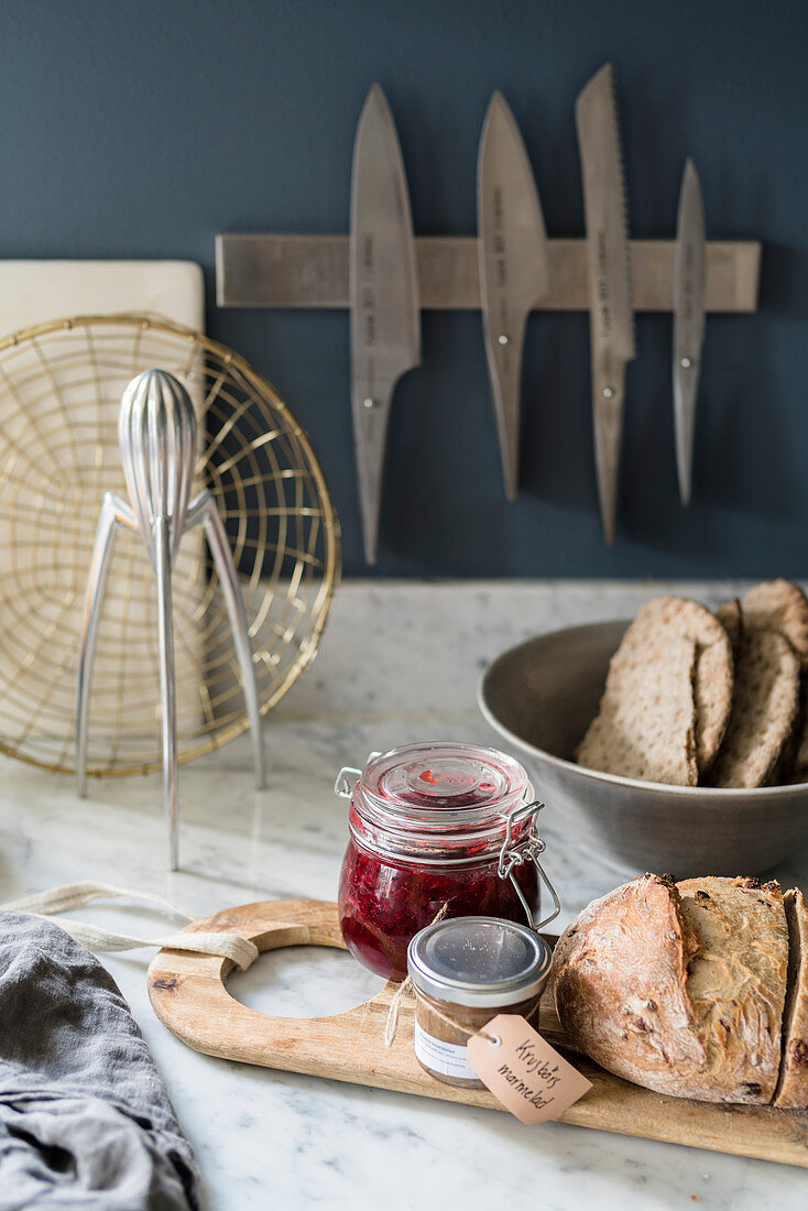 Holzbrett mit Marmeladen und Brot auf Marmor-Arbeitsplatte, im Hintergrund Messerhalter an blau-grauer Wand