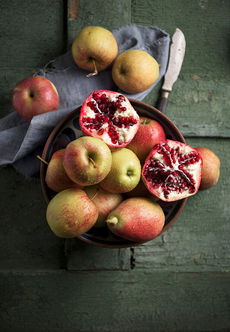 Äpfel, Birnen und Granatapfel in Schale auf grünem Holzuntergrund