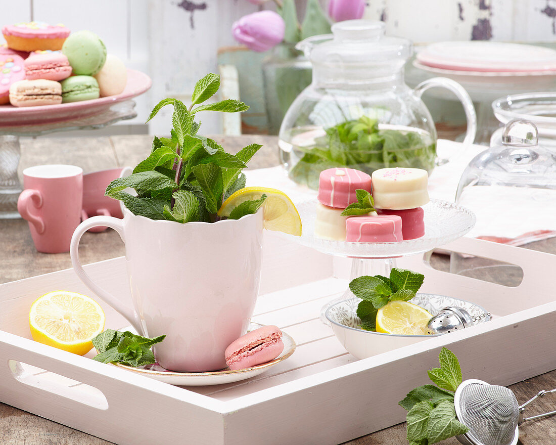 A cup of mint tea petit fours on a glass cake stand on a wooden tray