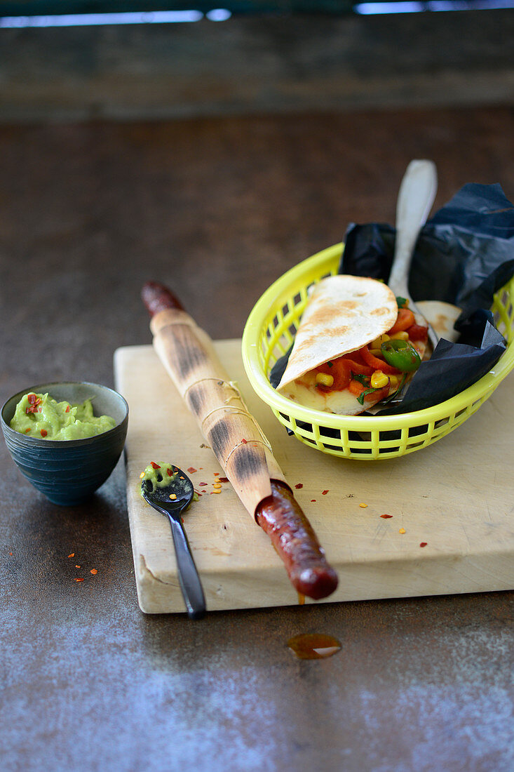 A chilli sausage wrapped in a wooden leaf with vegetables tacos and guacamole