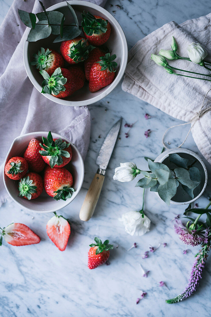 Bowls with ripe strawberries and beautiful flowers placed on white marble tabletop near knife