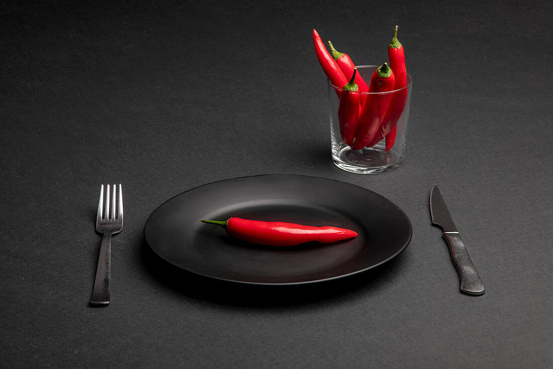 Fork and knife placed on black background near plate and cup with fresh chili peppers as representation of spicy dinner