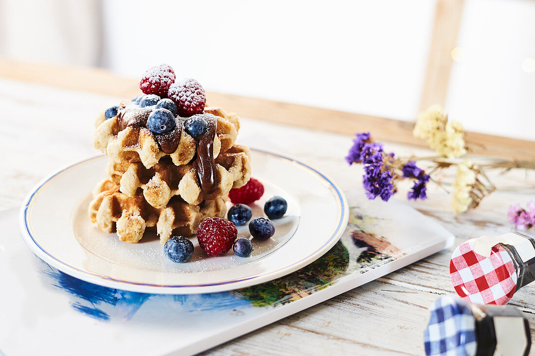 Plate with stack of golden waffles garnished with fresh berries and chocolate topping on table