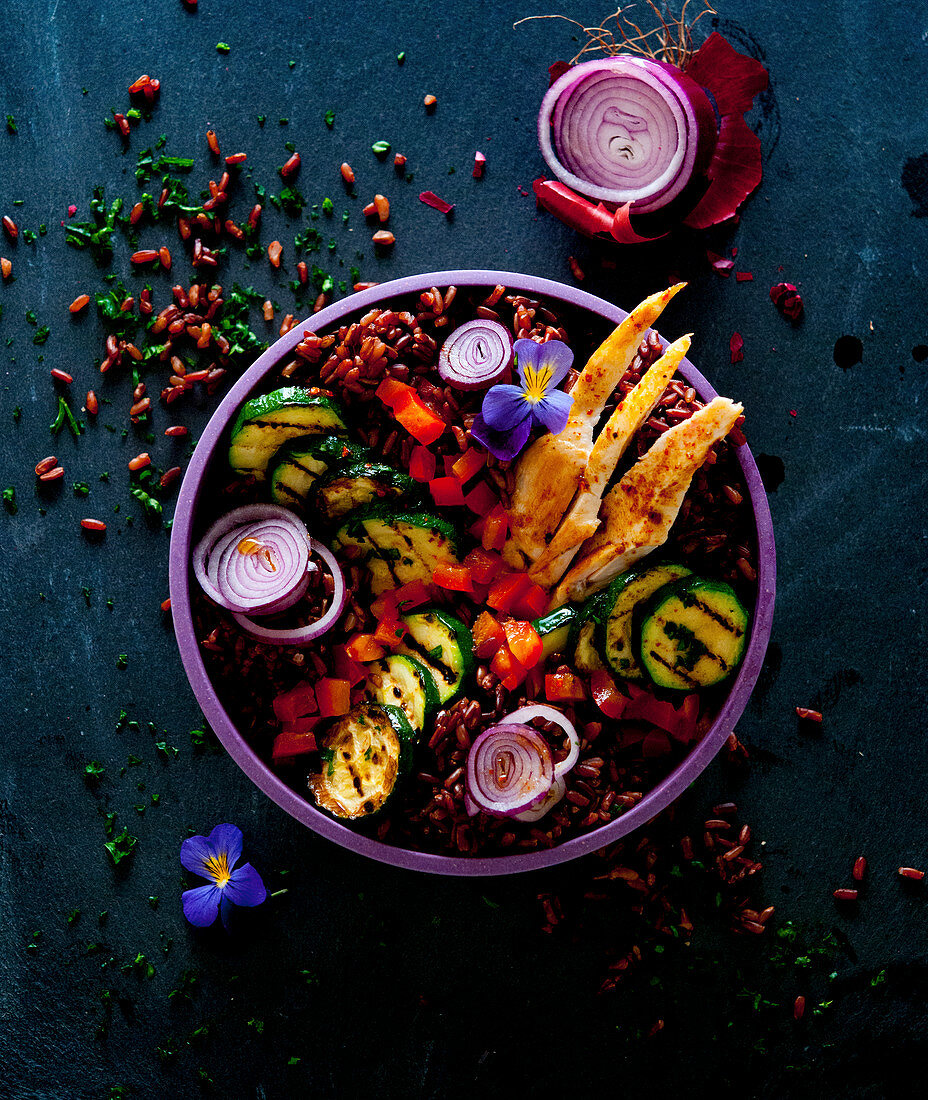 A bowl with red rice, pepper chicken, courgette and red onions