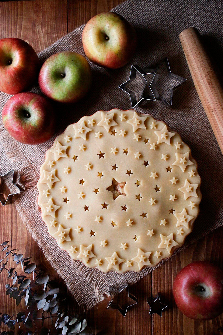 Star shaped cutters and rolling pin placed on wooden table near fresh apples and unbaked pie in kitchen