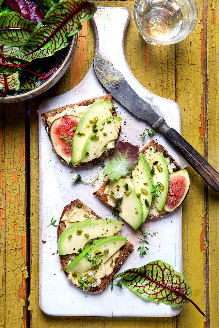 Slices of bread topped with avocado, figs, pistachio nuts, thyme and wild herb salad