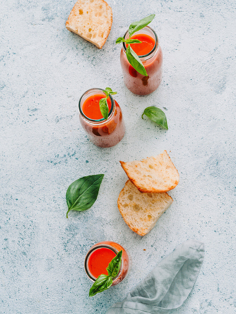 From above of glass bottles with fresh cold gazpacho soup on rough gray surface with toasts and basil