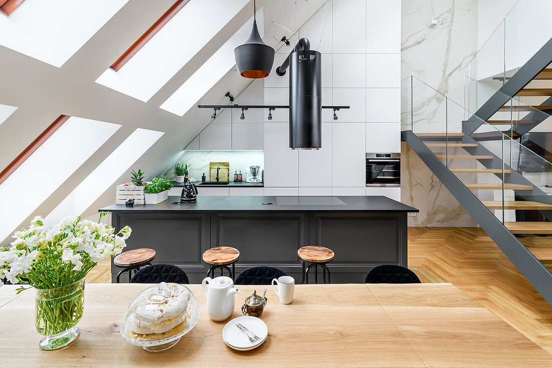 View across dining table to island counter and stairs in high-ceilinged room with skylights in sloping wall