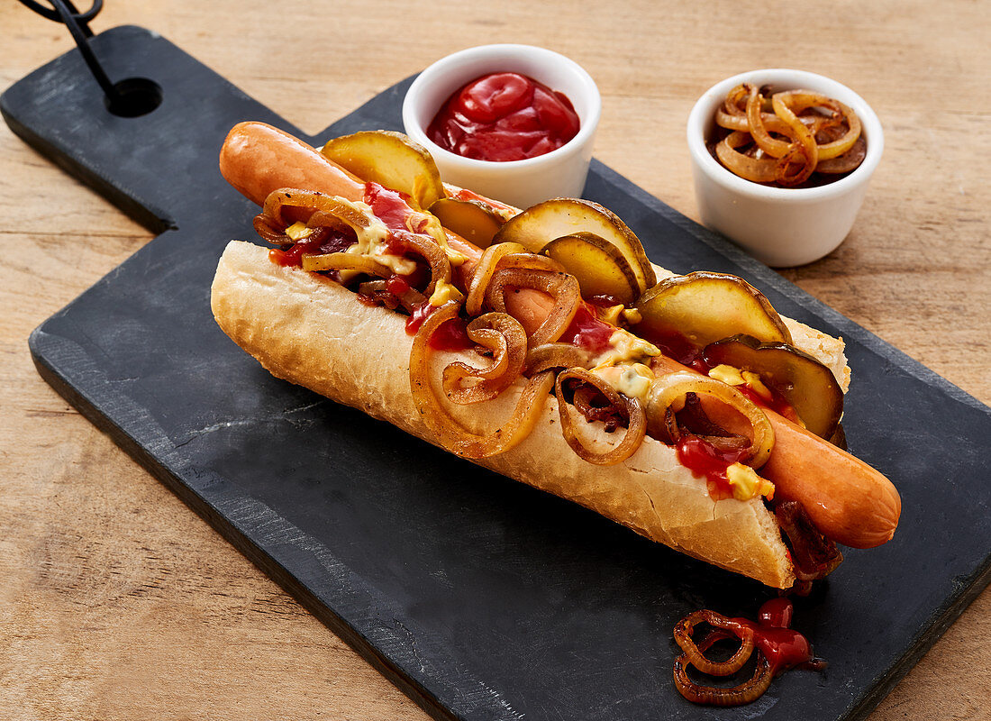 Hot Dog mit Röstzwiebeln, Essiggurken, Senf, Mayo und Ketchup