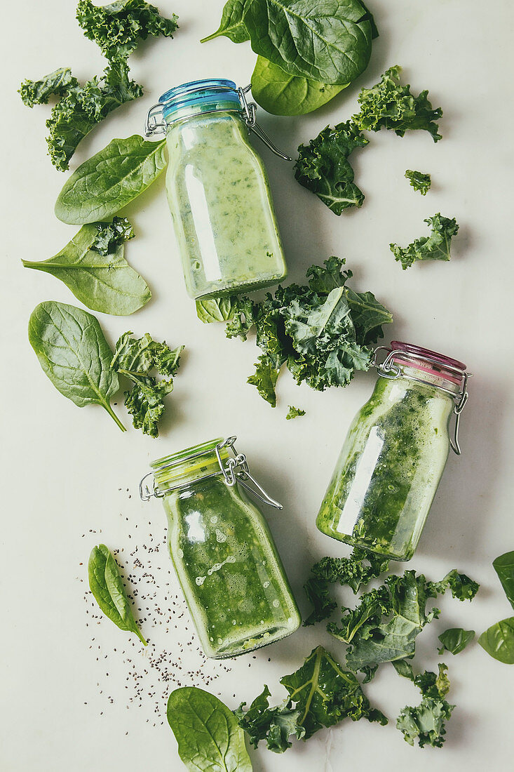 Variaty of green spinach kale apple smoothies in glass bottles with ingredients above over white marble background