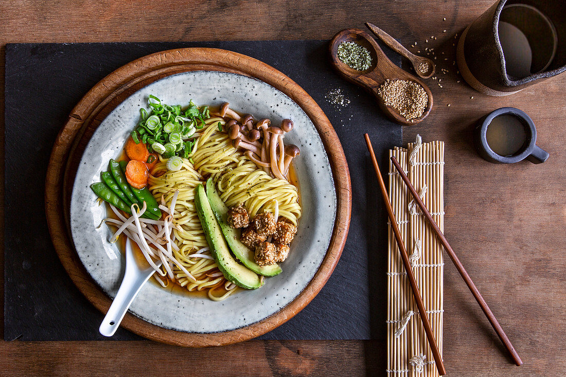 Vegan Japanese Ramen soup with avocado, sesame tofu and mushrooms in grey bowl over wooden background
