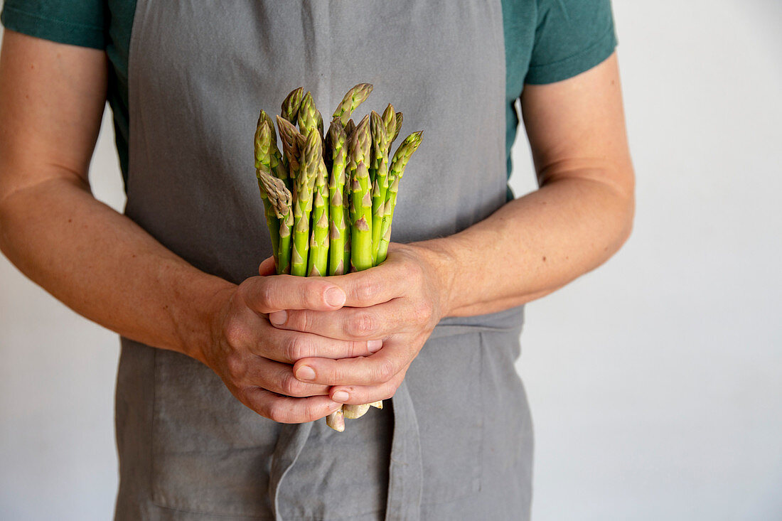 Man in grey apron before white background holding a bundle of green asparagus in his hands