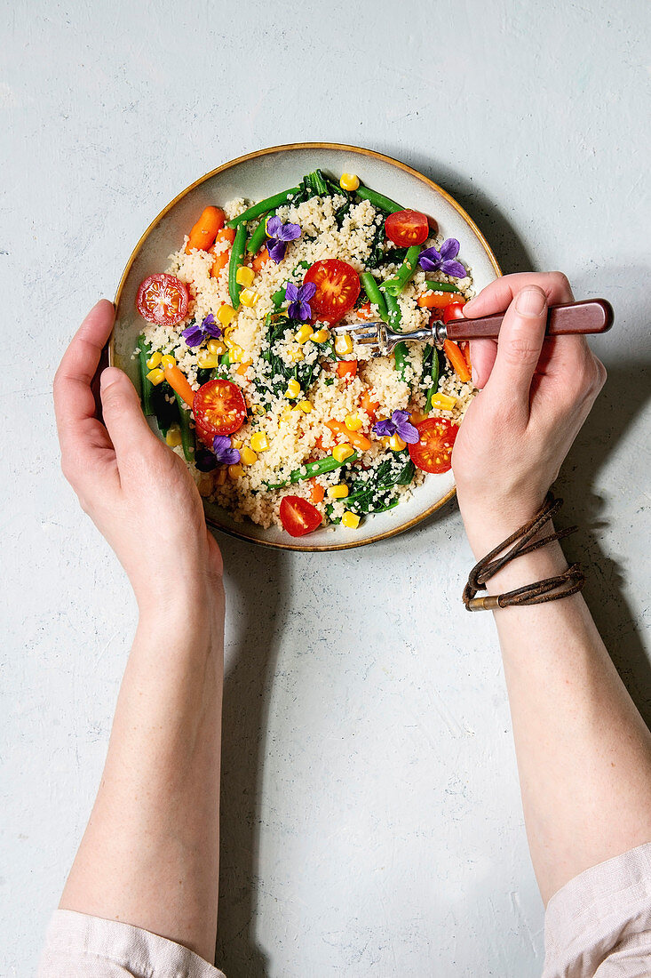 Couscous with parboiled vegetables baby carrots, green beans, sweet corn, spinach in ceramic plate with tomatoes, sesame and edible flowers