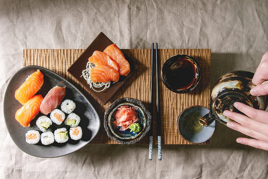 Sushi Set nigiri and sushi rolls on japanese wooden serving board with soy sauce, chopsticks, ceramic tea pot in hands