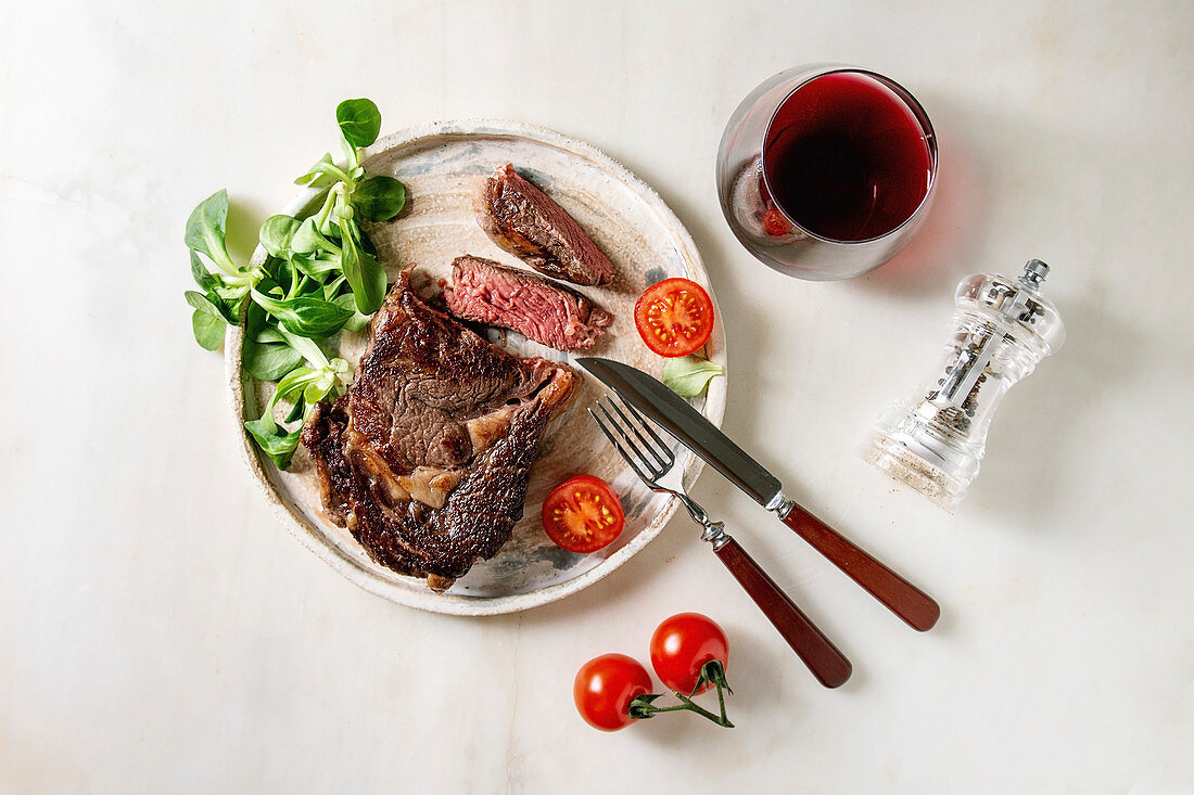 Grilled roasted sliced medium rare beef steak served in ceramic plate with green field salad, cherry tomatoes, cutlery, pepper, glass of red wine