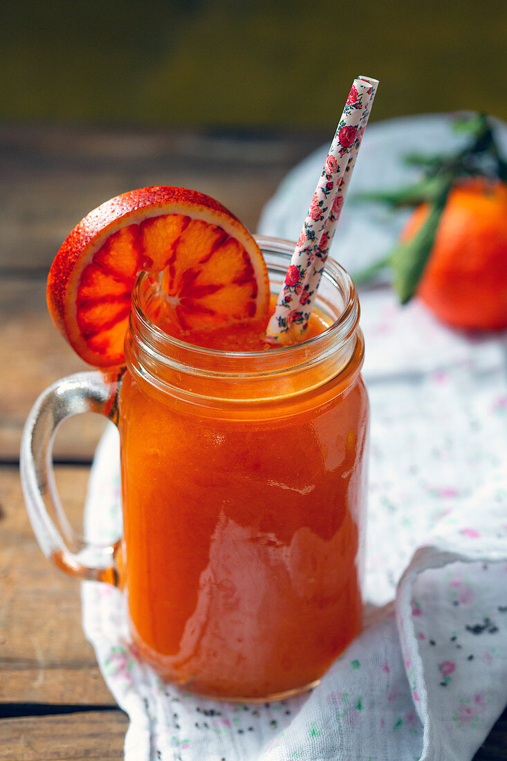 Jar of yummy fresh citrus juice with slice of ripe blood orange and straws placed on timber tabletop