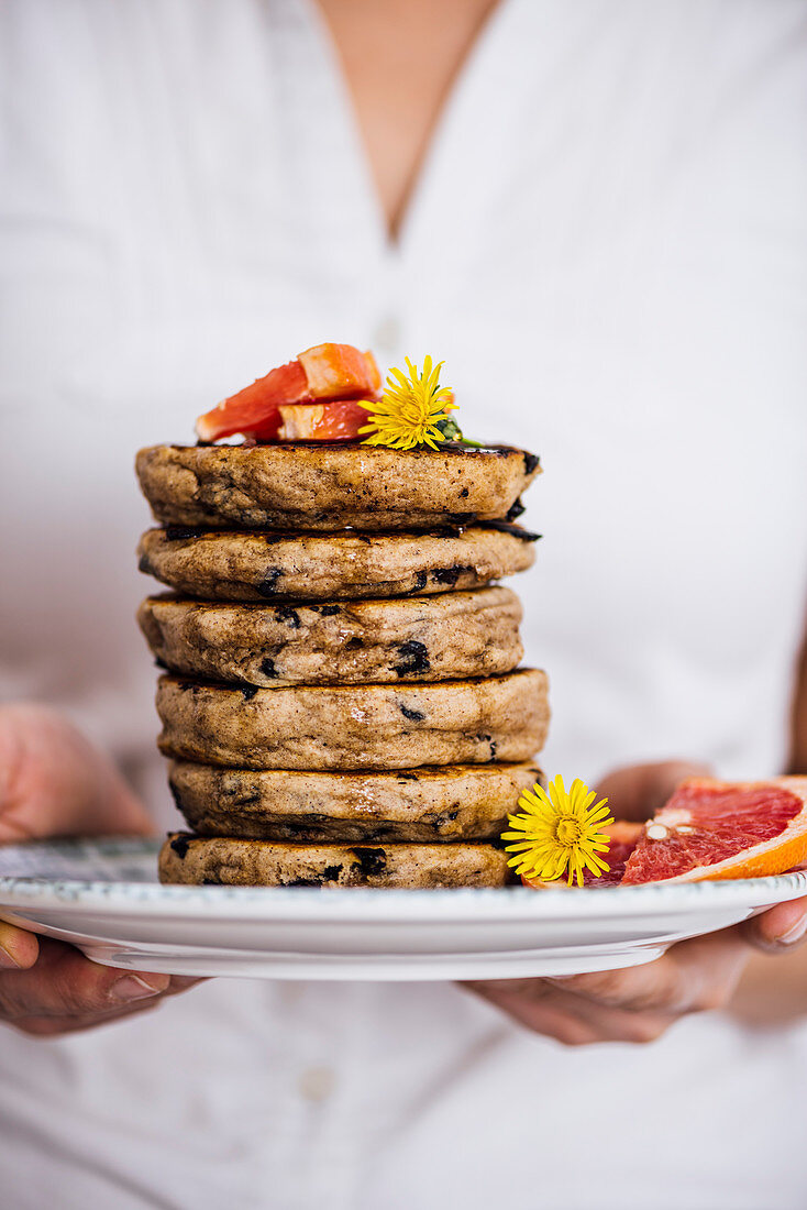 Woman holding a stack of pancakes with chocolate chips on a plate with flowers and grapefruit wedges