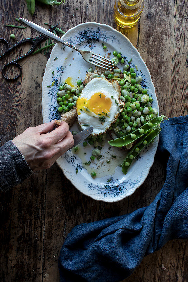 From above person hands eating bread toast with sauteed green peas and fried egg on wooden table