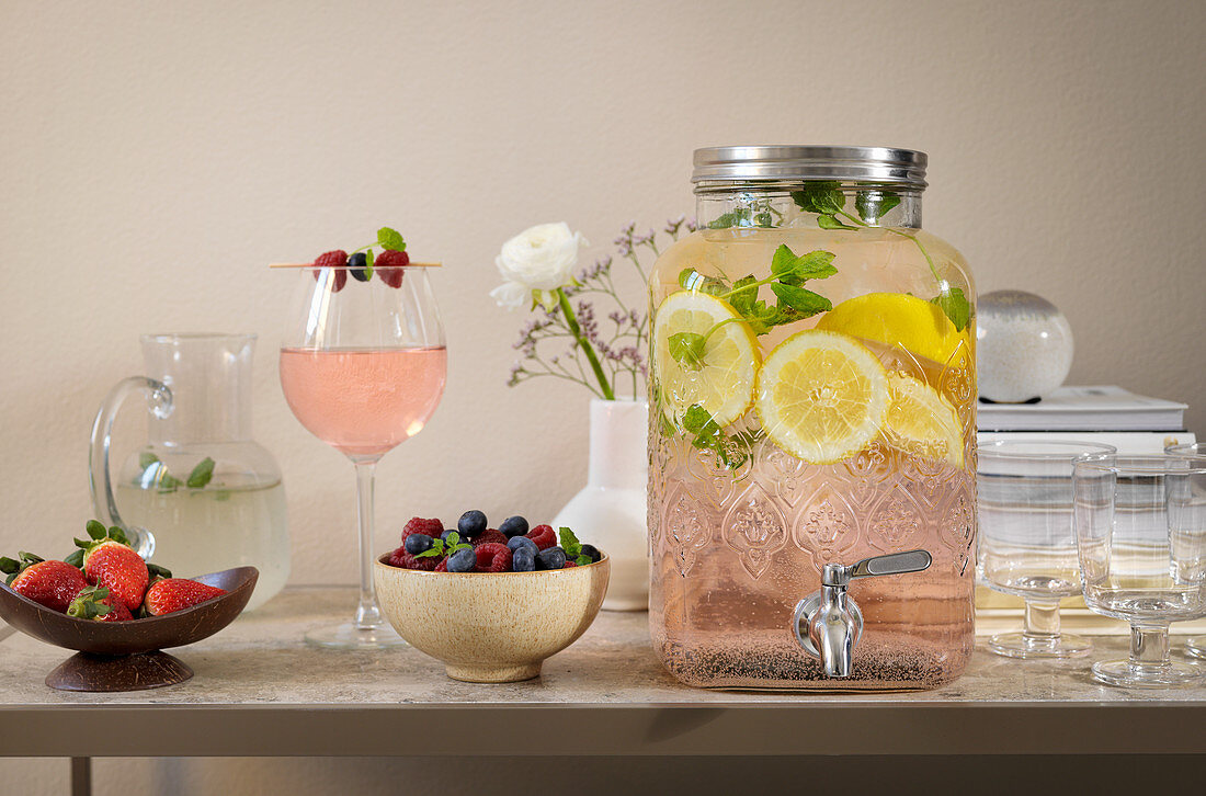 Erfrischungsgetränk mit Zitronenscheiben im Getränkespender, Beeren und Cocktail auf Tisch