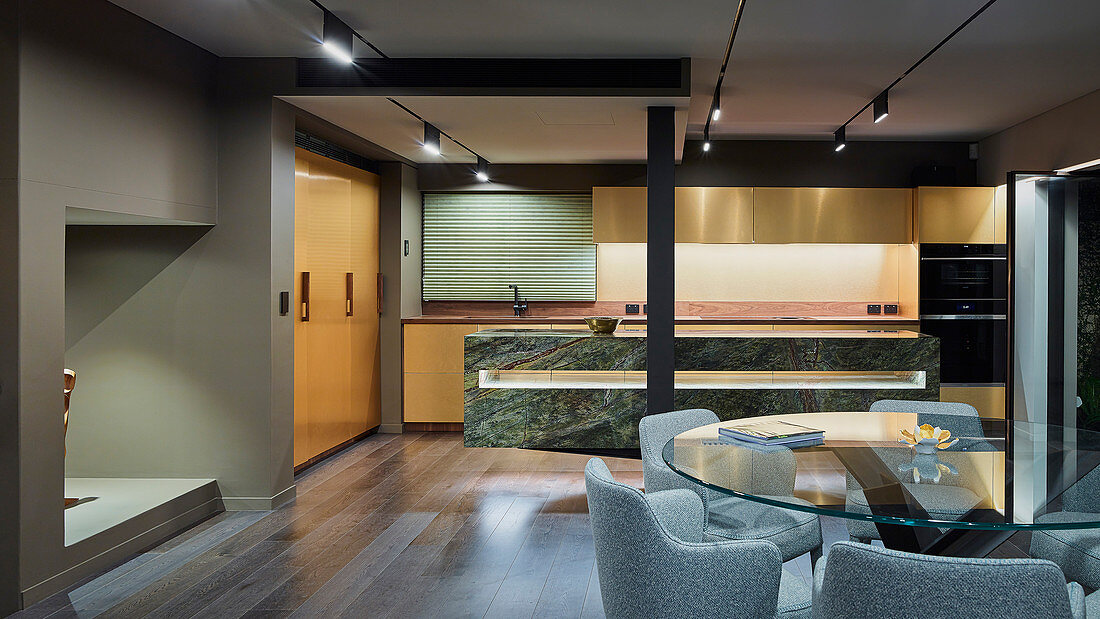 Round glass table, upholstered chairs and designer kitchen with marble counter in open-plan interior