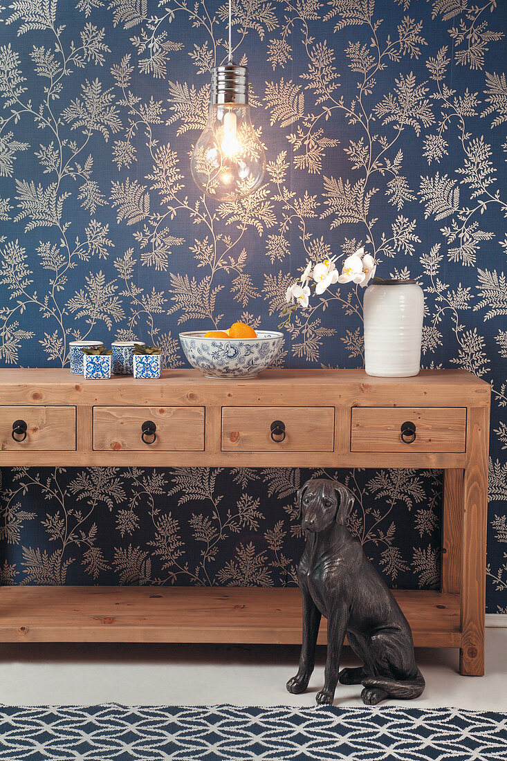 Wooden console table with drawers, light bulb lamp and dog sculpture against blue floral wallpaper